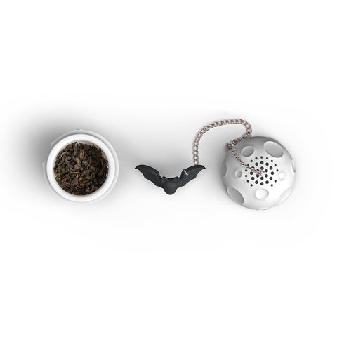 Loose tea in a cup beside the OTOTO Moonlit tea strainer with a bat-shaped charm on white background.