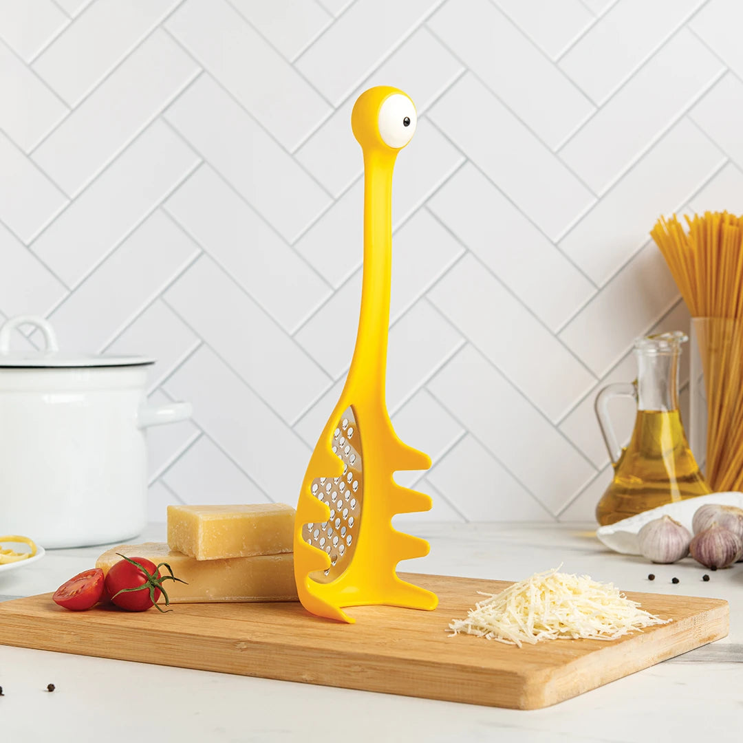 Multi Monster grater on a kitchen counter with Parmesan, tomatoes, spaghetti, and dinosaur design.