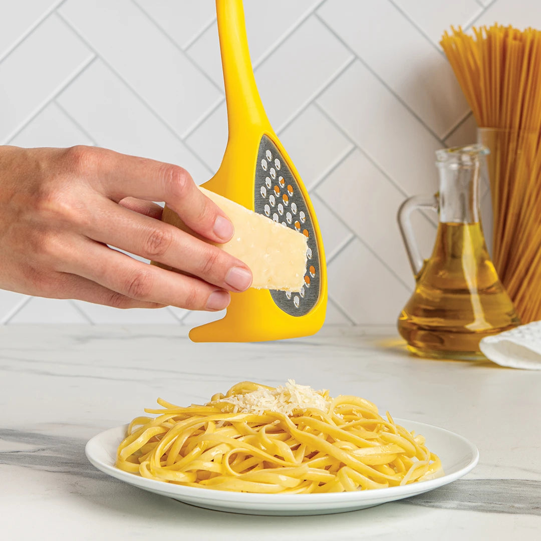 Grate cheese over pasta using the Multi Monster with oil and a pasta spoon in the background.