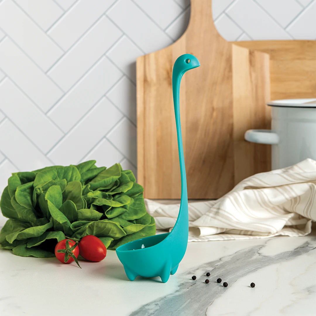 Nessies dinosaur-shaped ladle on a counter, surrounded by lettuce, tomatoes, and peppercorns.