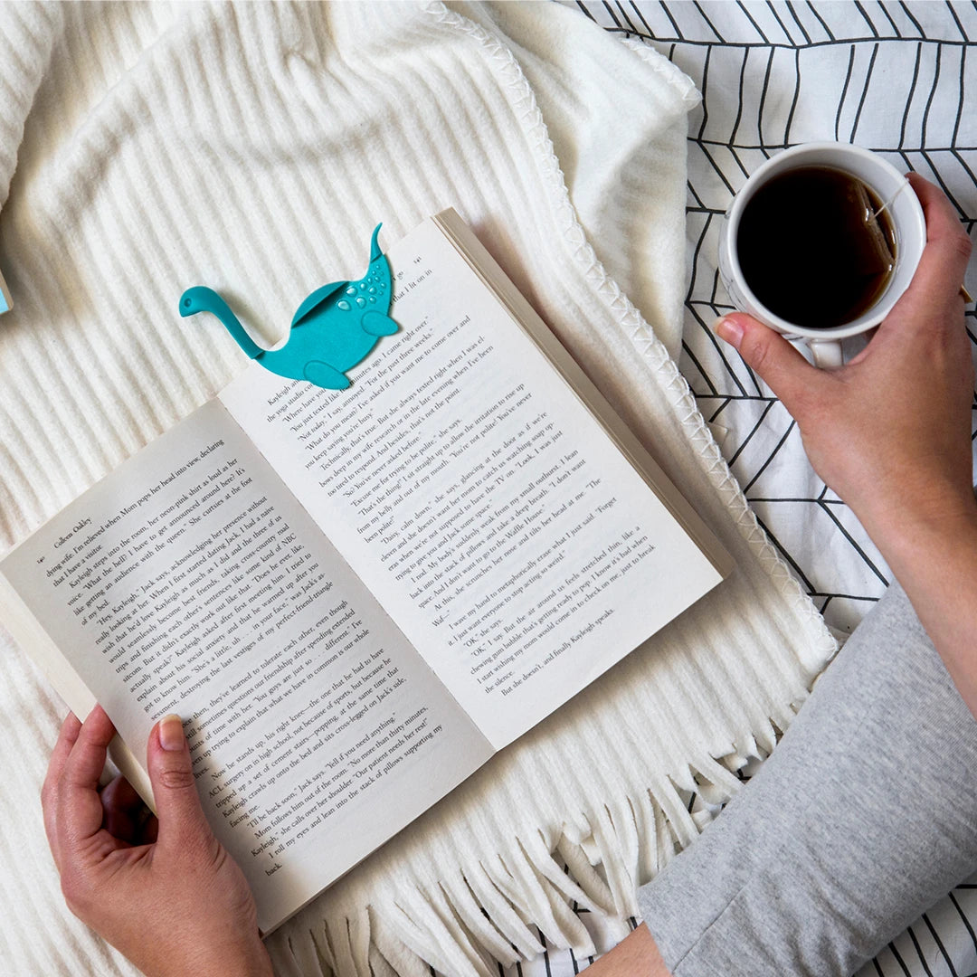 Reading a book with a Nessie Tale bookmark, holding coffee, on a cozy blanket.