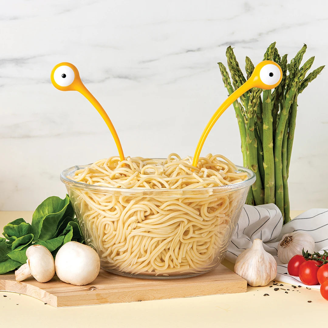 A bowl of noodles with quirky Pasta Monsters utensils, surrounded by fresh vegetables.