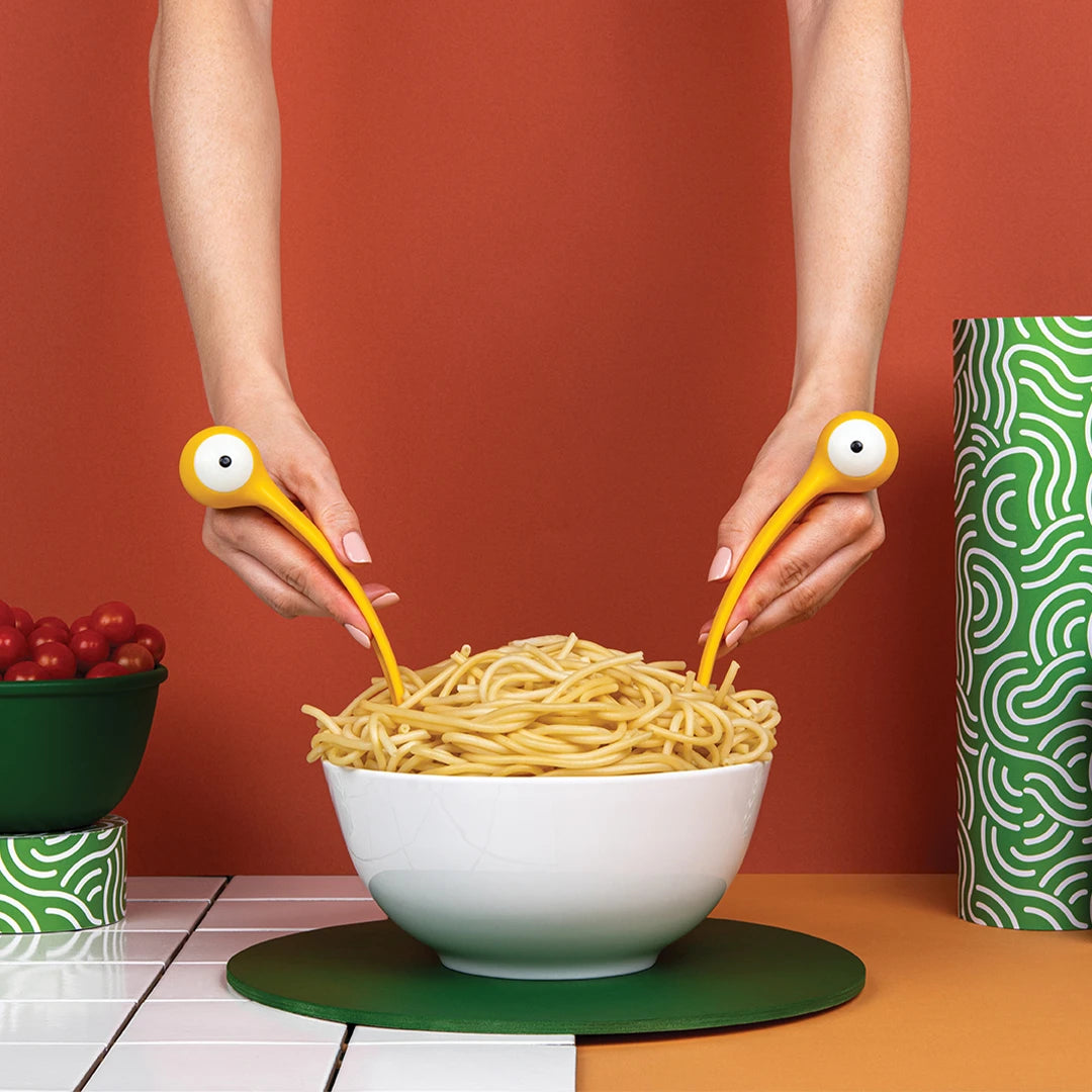 Person serves spaghetti from a white bowl on a colorful table using Pasta Monsters utensils.