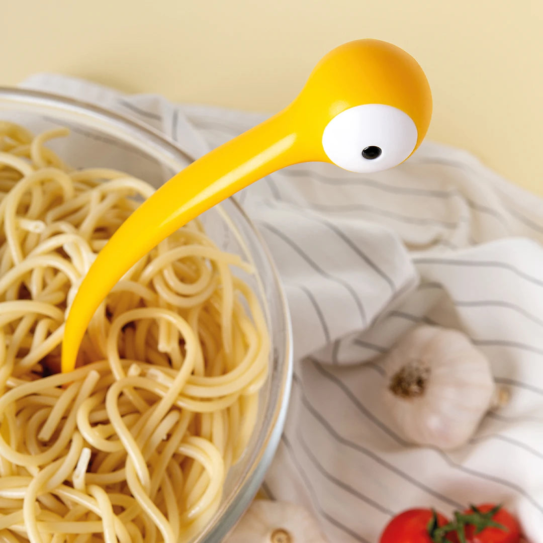 A yellow Pasta Monsters ladle with an eye rests in spaghetti, surrounded by garlic and tomatoes.