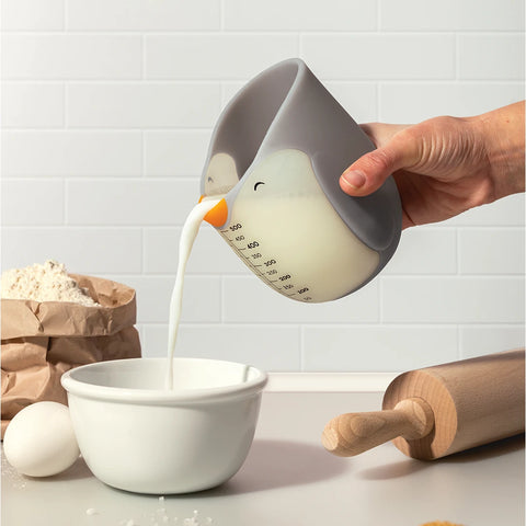 A hand pours milk from OTOTO's Penny, a cute penguin-shaped measuring cup, with baking items nearby.