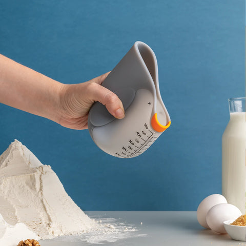 A hand squeezes an OTOTO Penny cute measuring cup near flour, eggs, and milk on a blue background.