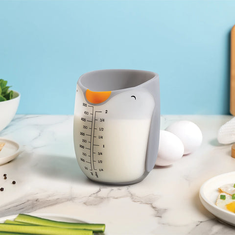 A cute OTOTO Penny penguin-shaped measuring cup filled with milk sits on a kitchen counter.