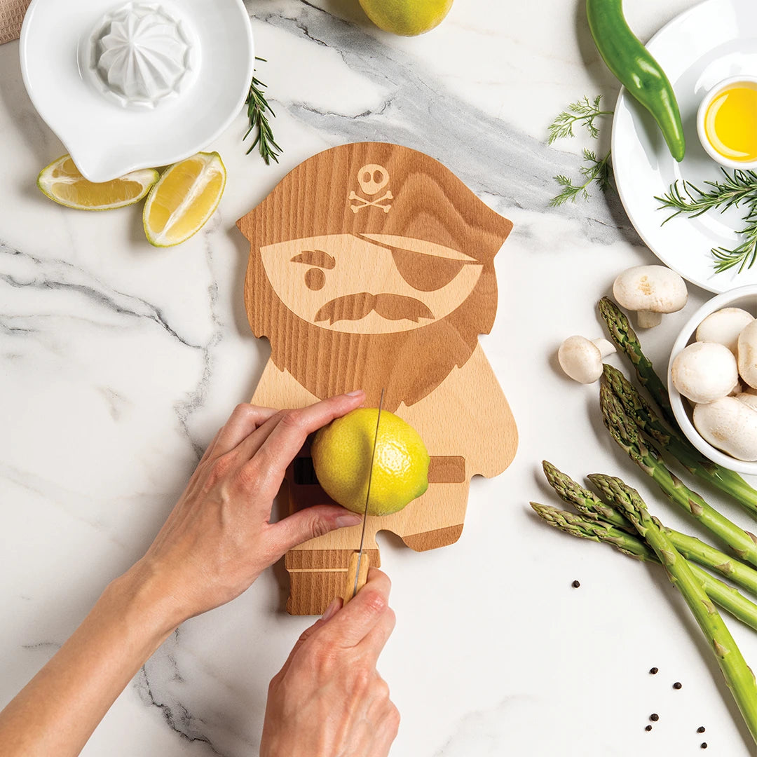 Hands slicing lemon on a pirate-shaped cutting board, surrounded by various vegetables and ingredients.