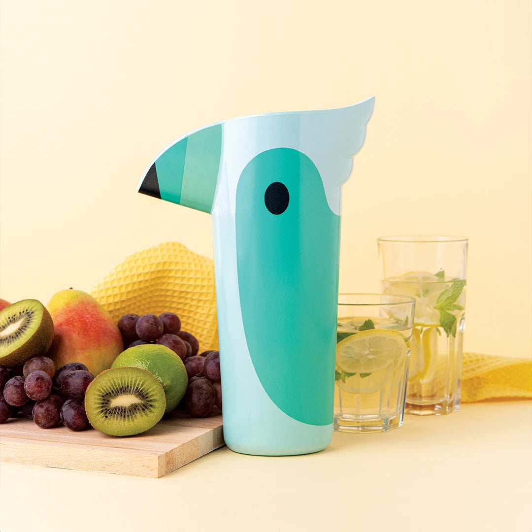 Polly pitcher with teal bird design, glasses, and towel on wooden board with fruits.