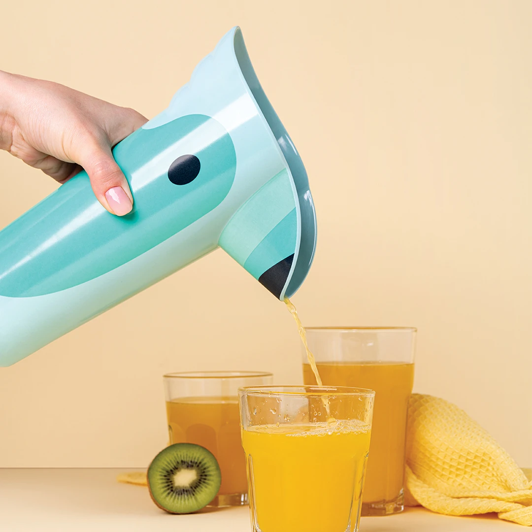 Pouring juice from a Polly into a glass, next to kiwi and another full glass, on a yellow background.