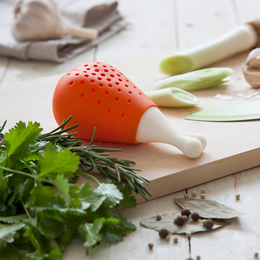 OTOTOs Pulke herb infuser, shaped like a chicken drumstick, sits on a board with fresh herbs and veggies.