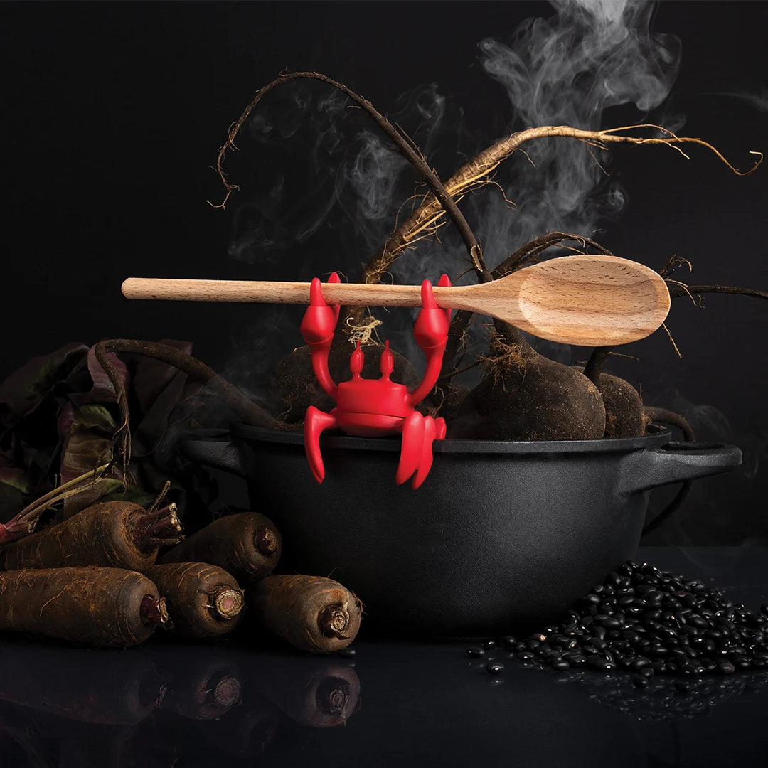 OTOTOs Red silicone crab grips a wooden spoon, surrounded by dark veggies and beans.