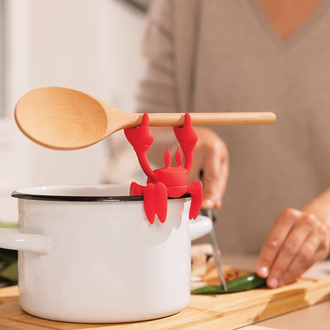 Red OTOTO silicone utensil rest on pot; person chopping veggies nearby.