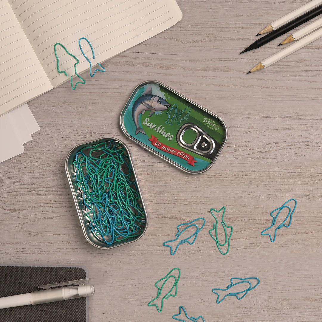 The Sardine Paper Clips by OTOTO sit on a desk, their sardine can-shaped case flanked by pencils and a notebook.