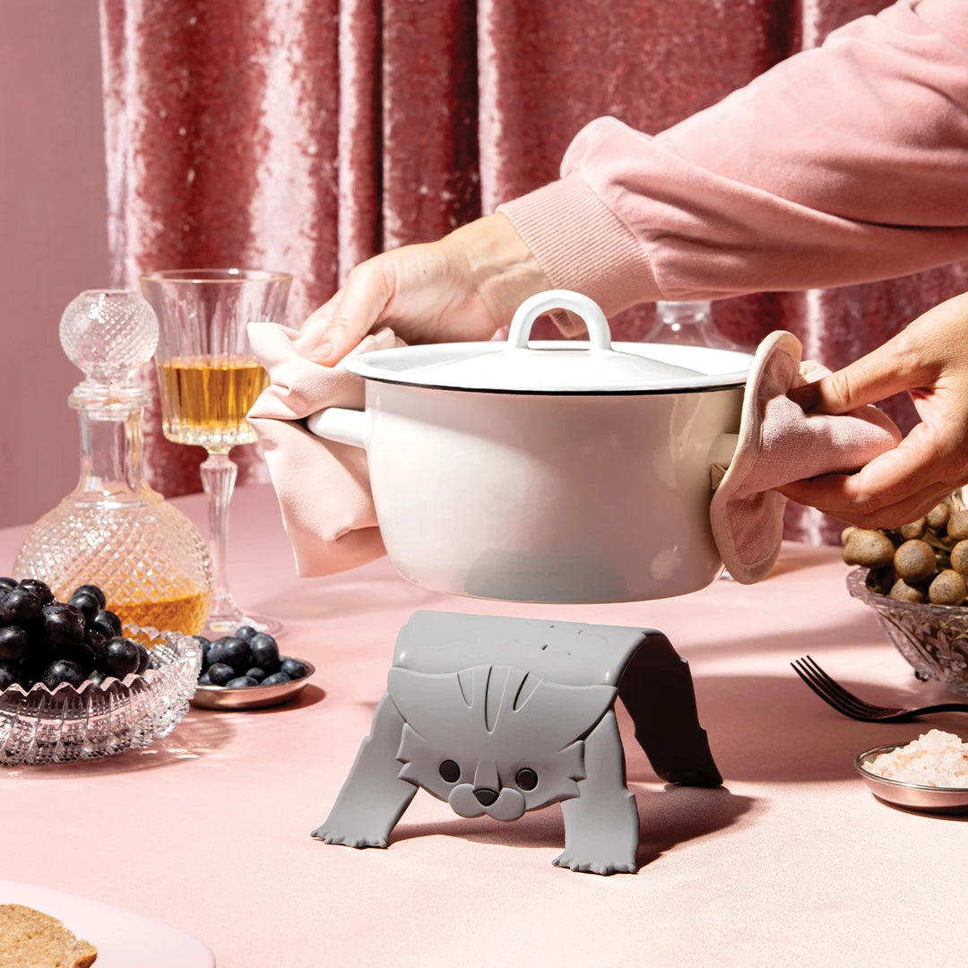 A pot placed on the Scaredy Cat trivet, surrounded by elegant dinnerware on a pink tablecloth.