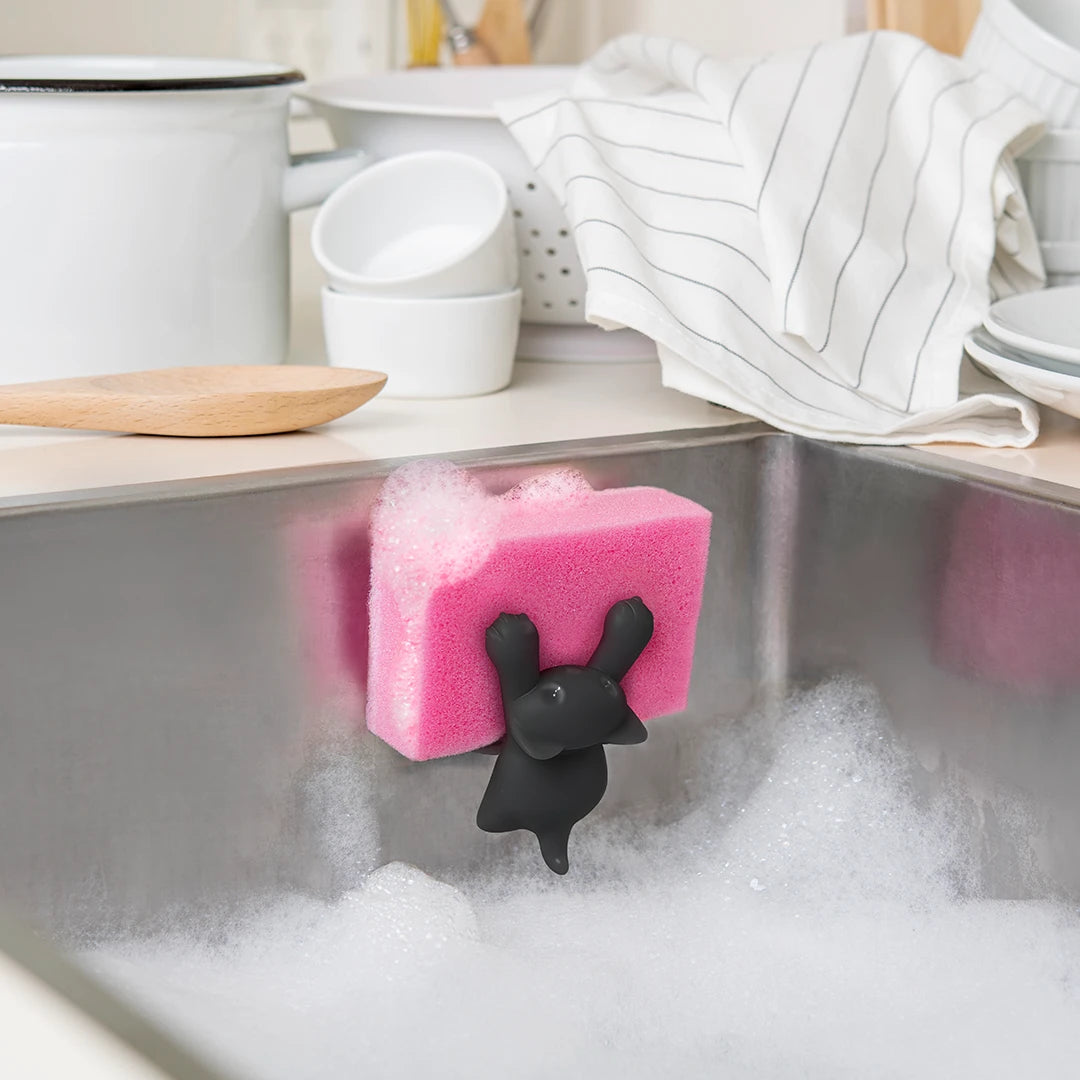 A pink Scrubby with a cat holder clings to a sink filled with soapy water.