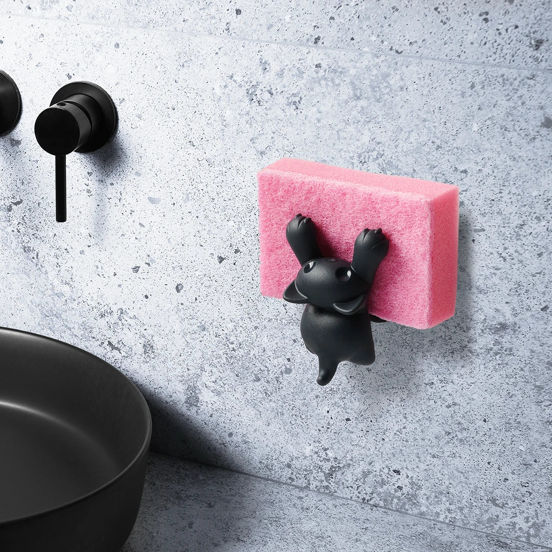 Scrubby by OTOTO: black cat sponge holder with pink scrubby on gray tiled wall near the sink.