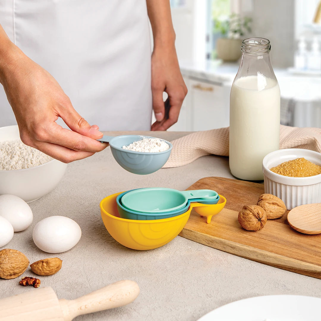 Person using a blue measuring cup in a kitchen with flour, milk, eggs, and other ingredients.
