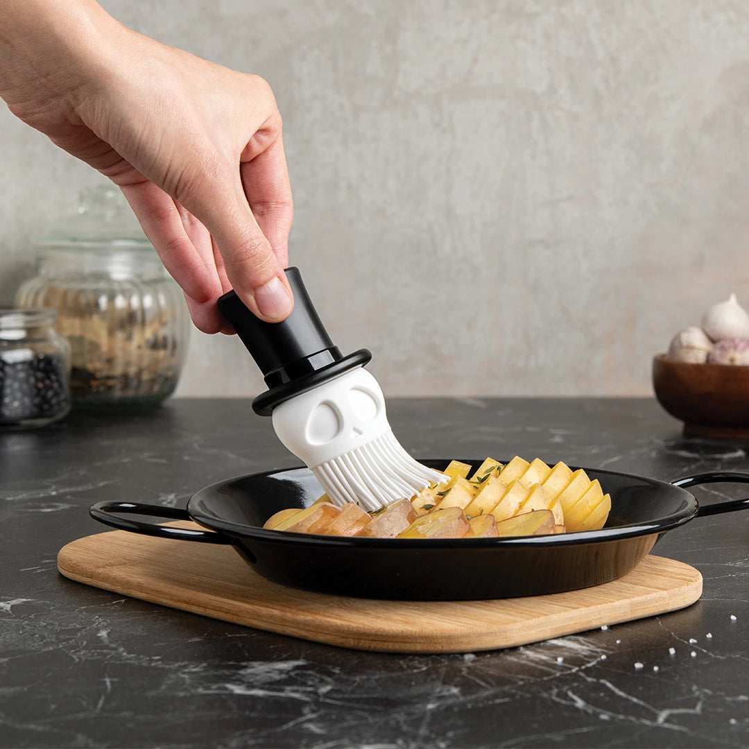 A Skully basting brush is used on sliced potatoes in a black pan for cooking adventures.