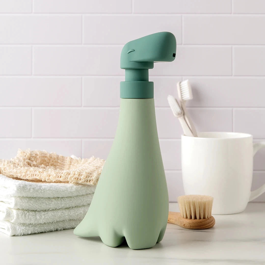 Soapasaurus hand soap dispenser on a bathroom counter among towels, toothbrushes, and a cup.