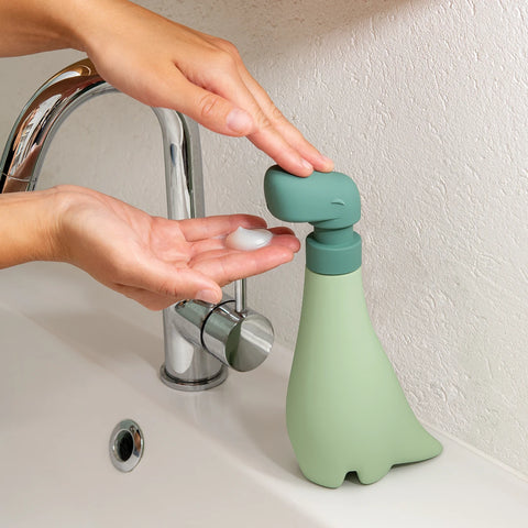 Person using the Soapasaurous hand soap dispenser by a sink.
