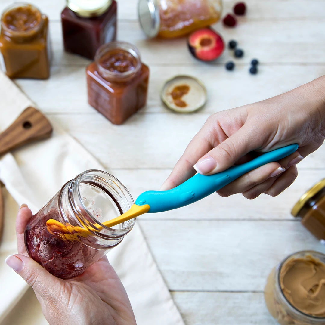 Person using a blue Splatypus to spread jam from a jar, with other jars on a wooden table.