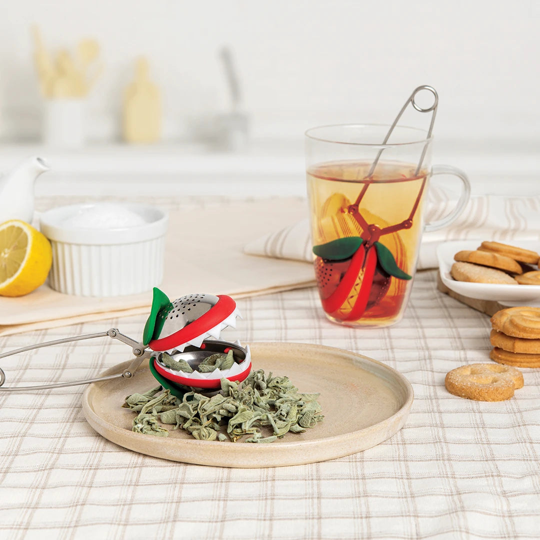 Tea infuser shaped like a Venus flytrap next to a mug and plate of herbs on a table with cookies.