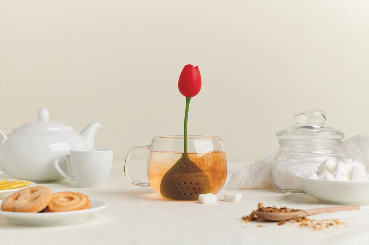Glass cup with Tulip infuser by OTOTO, surrounded by teapot, cookies, and sugar.