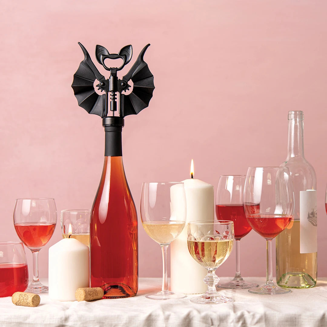 Wine glasses, bottles, candles, and an OTOTO Vino bat-shaped corkscrew on a pink background table.