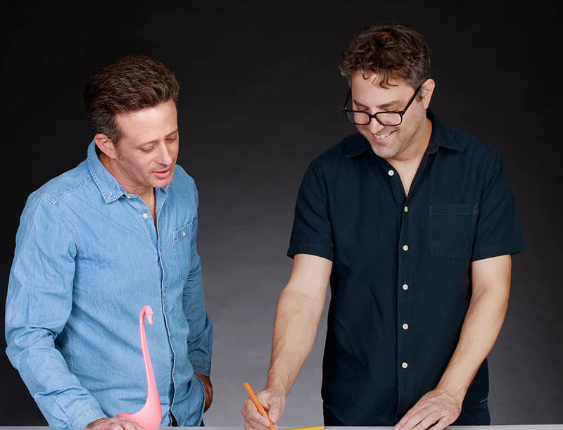 Two men collaborate over papers, with a pink flamingo figurine on the table.