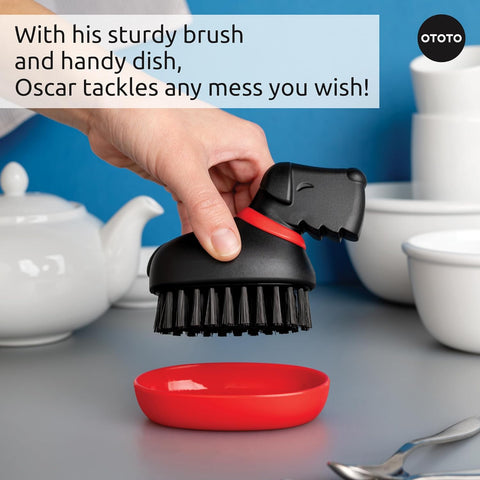 A hand holds the OTOTO Oscar dog-shaped dish brush over a red plate, with bowls in the background.