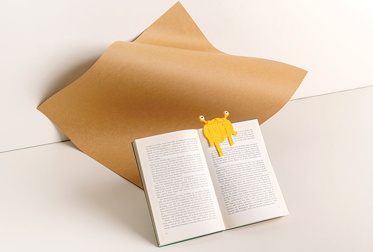 Open book with a quirky yellow Spaghetti Tale bookmark beside a large sheet of brown paper on a white background.