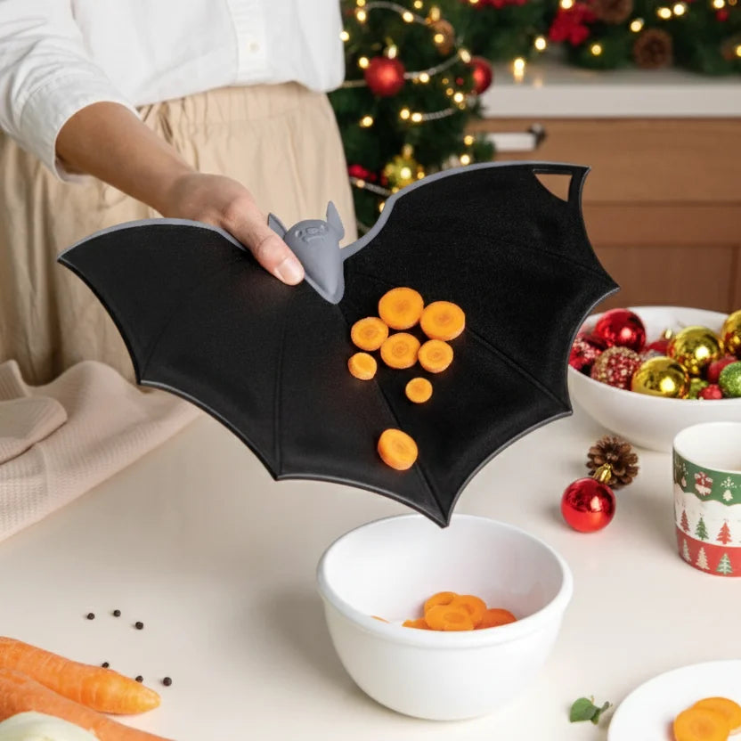 Someone slices carrots with Batty, a bat-shaped cutter, on a plastic board in a festive kitchen.