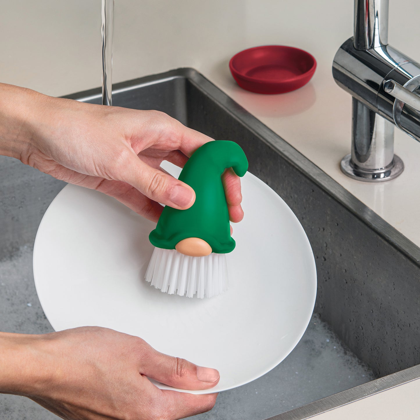A person washes a plate with the OTOTO Beardy gnome dish brush by the kitchen sink.