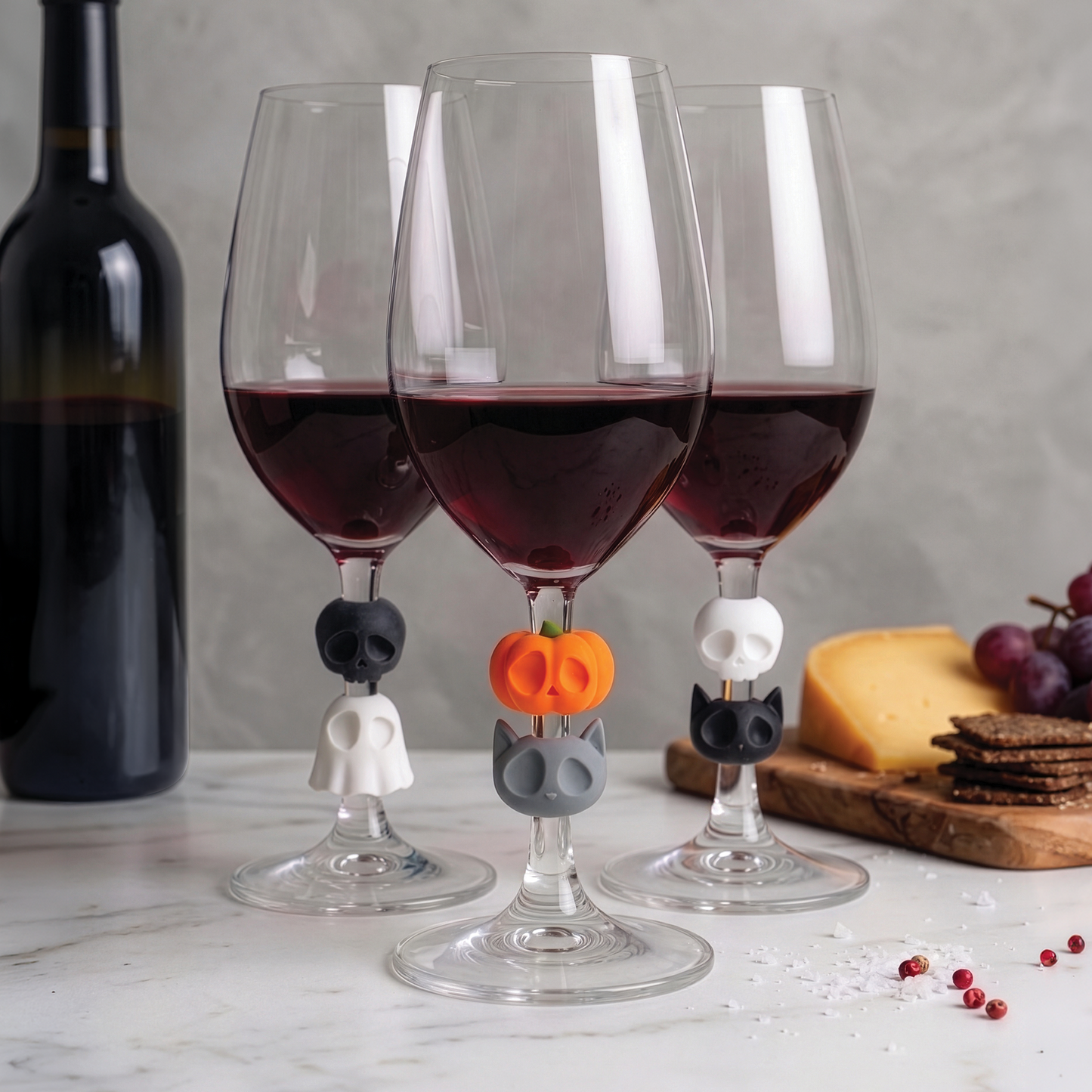 Three Boo Team glasses with skull and pumpkin charms, filled with red wine, beside cheese and grapes.