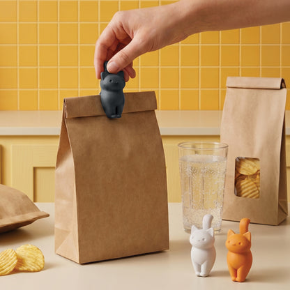 A hand seals a paper bag with a CatSnap; two CatSnaps and snacks sit on the counter.
