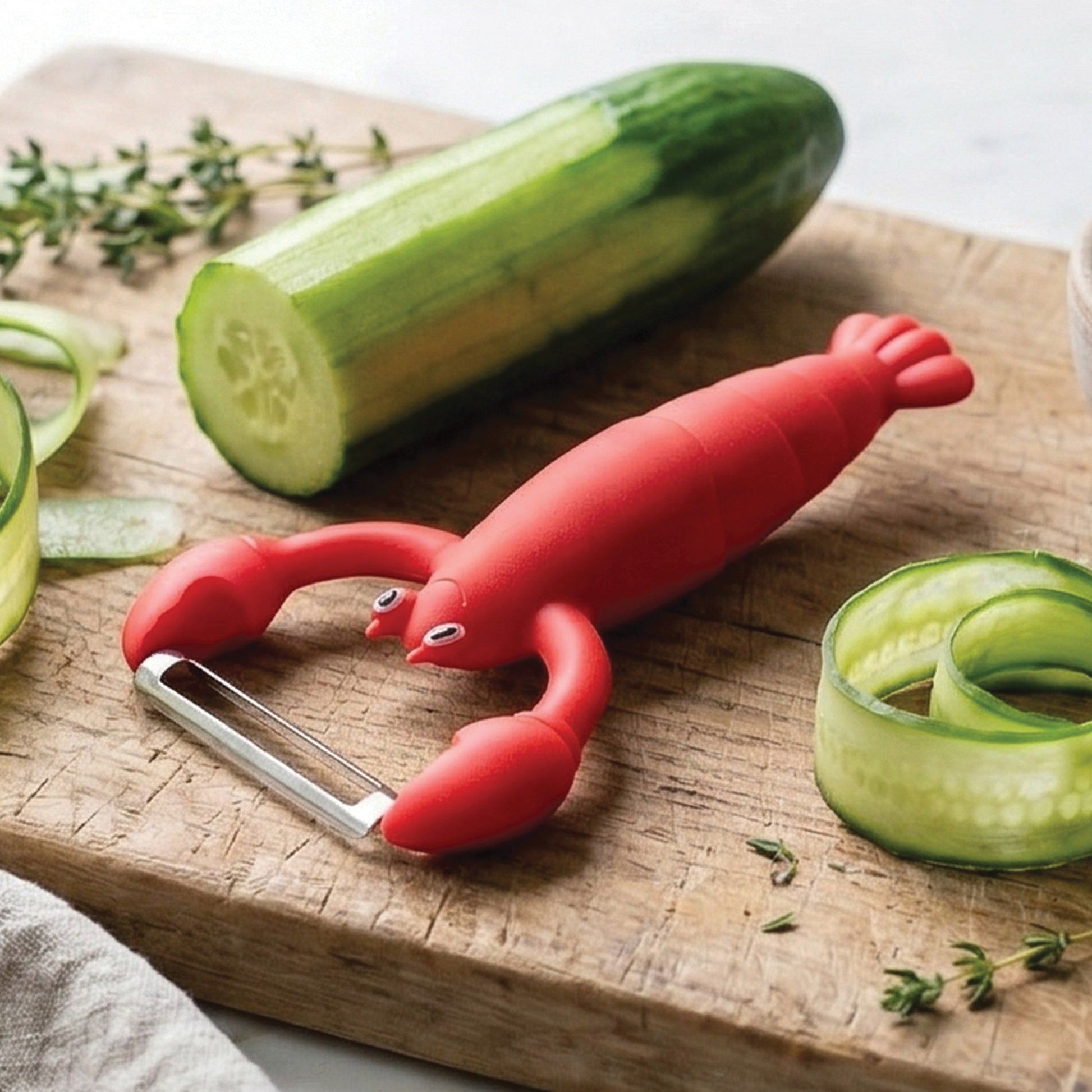 The OTOTO Clawy lobster-shaped vegetable peeler sits by a cucumber on a cutting board.