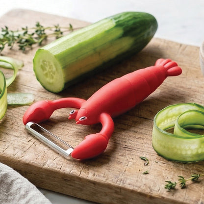 The OTOTO Clawy lobster-shaped vegetable peeler sits by a cucumber on a cutting board.