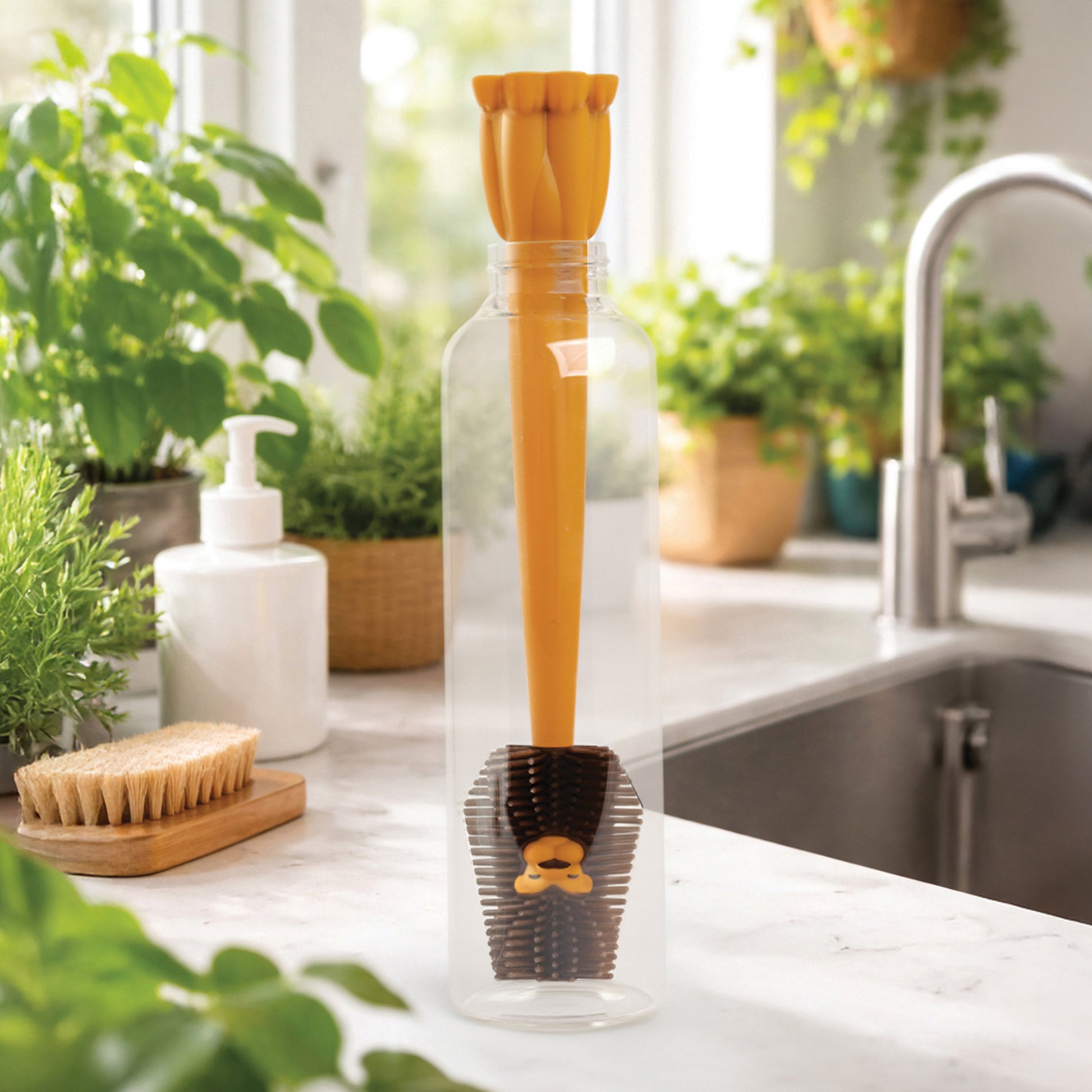 A yellow OTOTO Cleo bottle cleaner stands in a clear bottle on a kitchen counter with plants nearby.