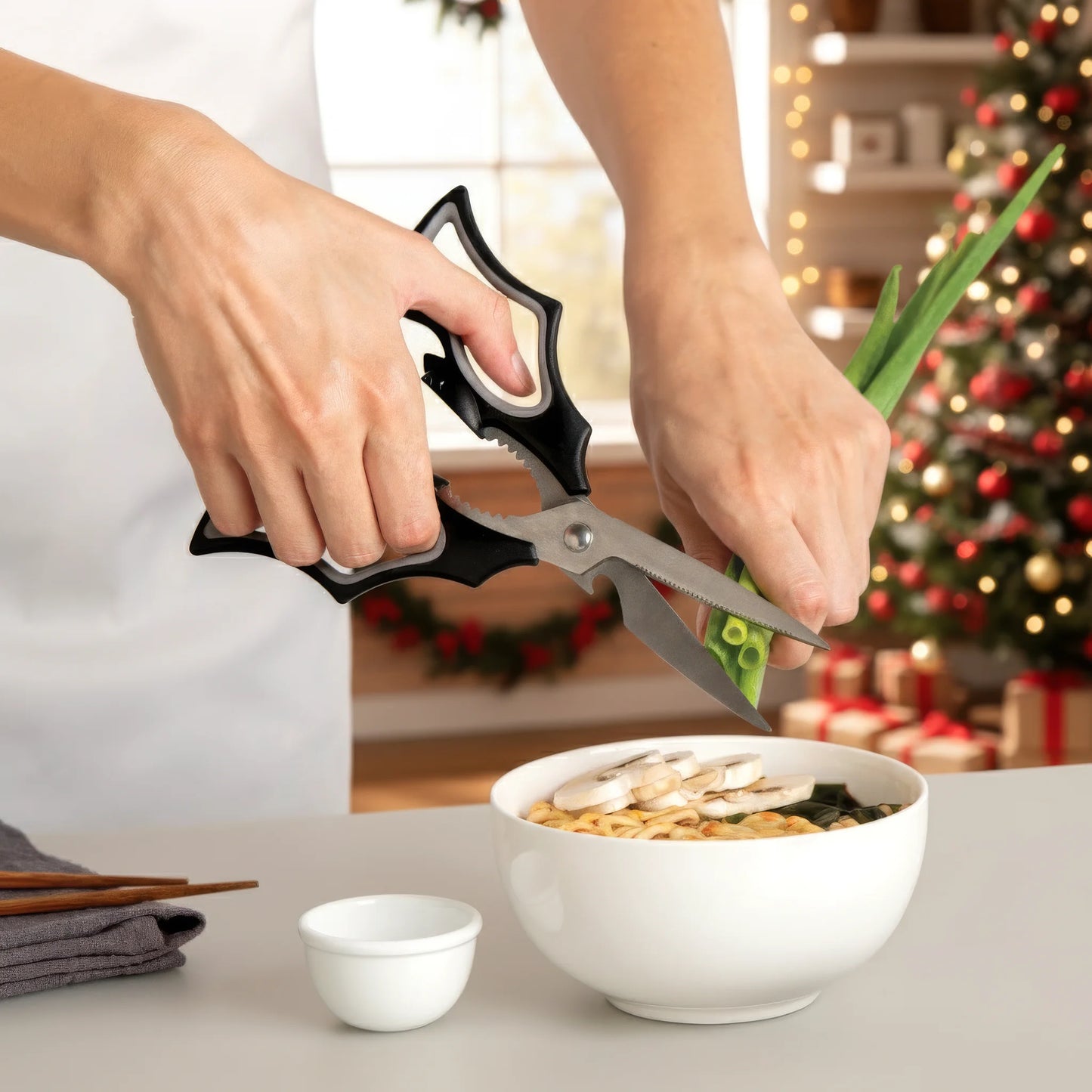 Hands using Elizabat to cut green onions over noodles, with a Christmas tree in the background.