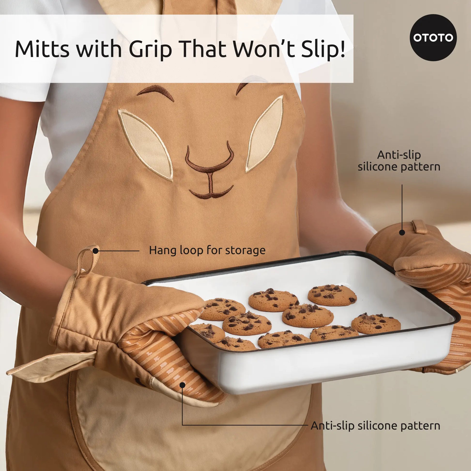 Person wearing Aproo bunny kitchen apron and oven mitts, holding a tray of chocolate chip cookies.