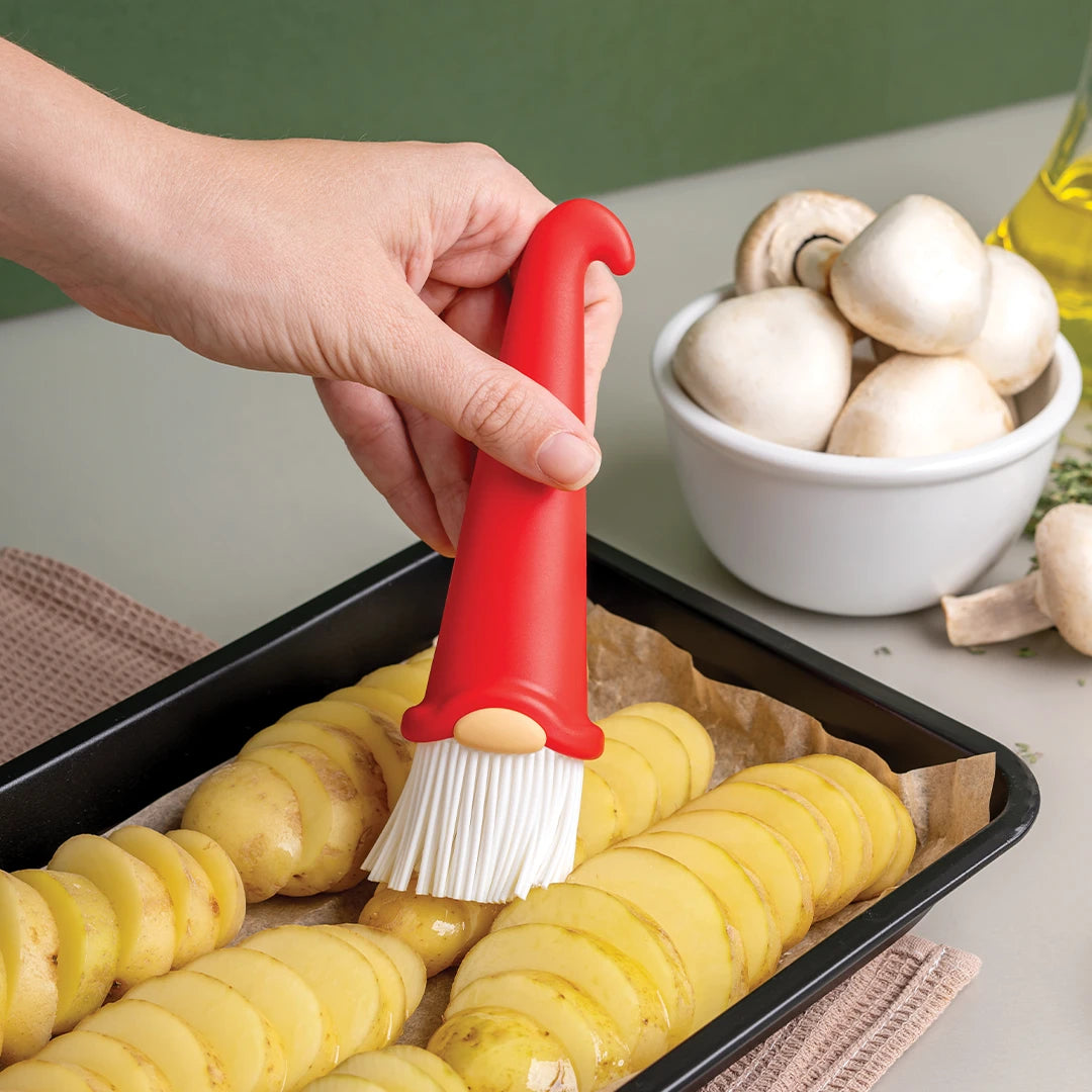 A hand uses Gnomeo, a red gnome-shaped brush by OTOTO, to coat sliced potatoes in a baking tray.