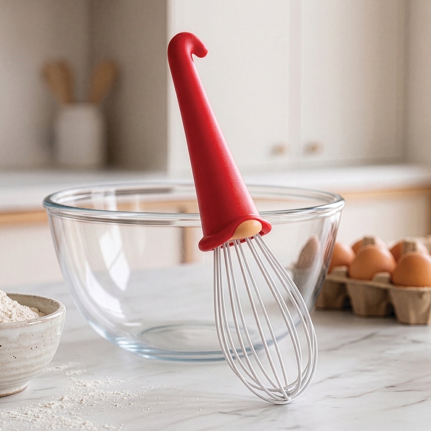 A Gnomeo whisk with a red handle rests in a glass bowl, surrounded by eggs and flour.