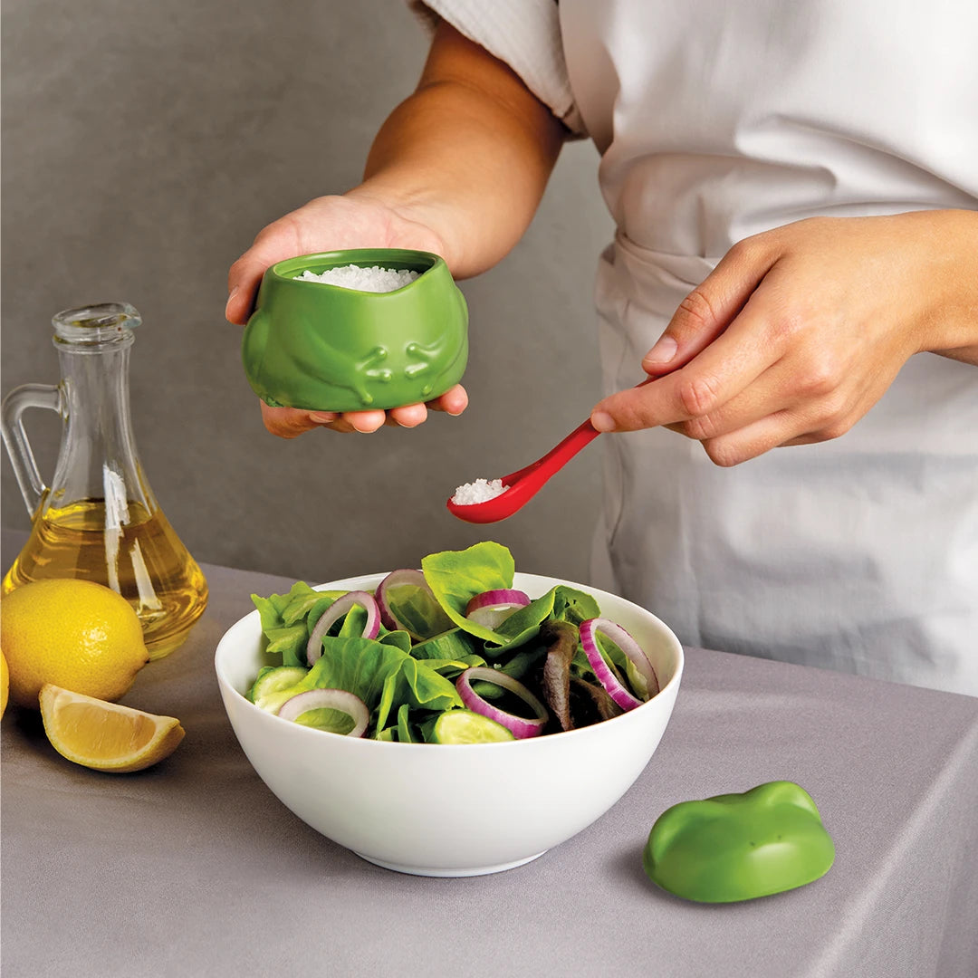 Lily-shaped container among lemons and oil as someone seasons a salad.
