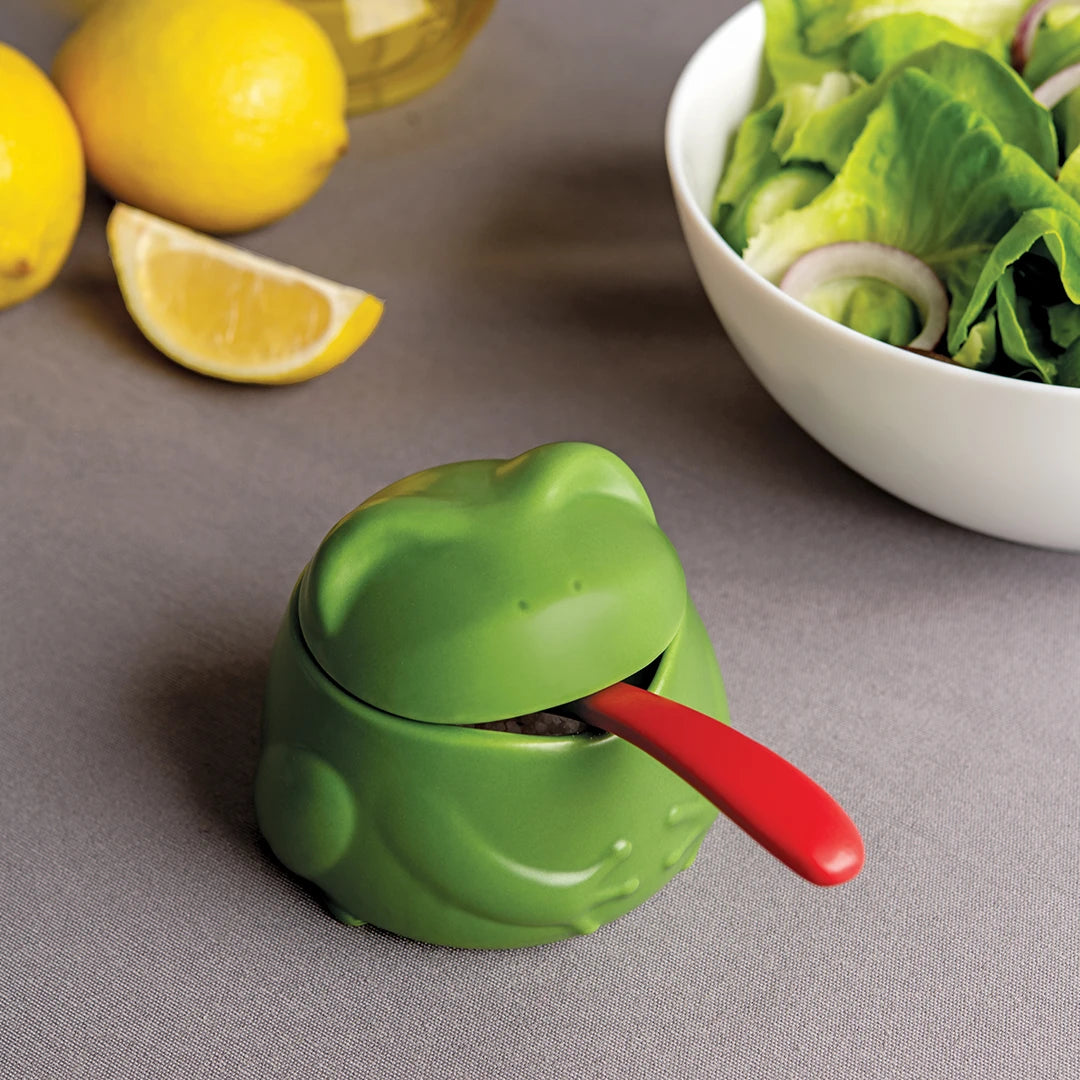 Lily frog container with red spoon by lemons and salad on gray surface.