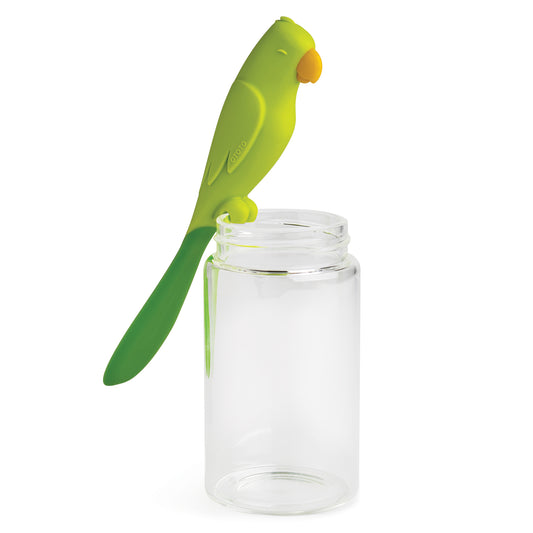 Parry, the green parrot-shaped silicone spatula by OTOTO, sits on a clear, empty glass jar.