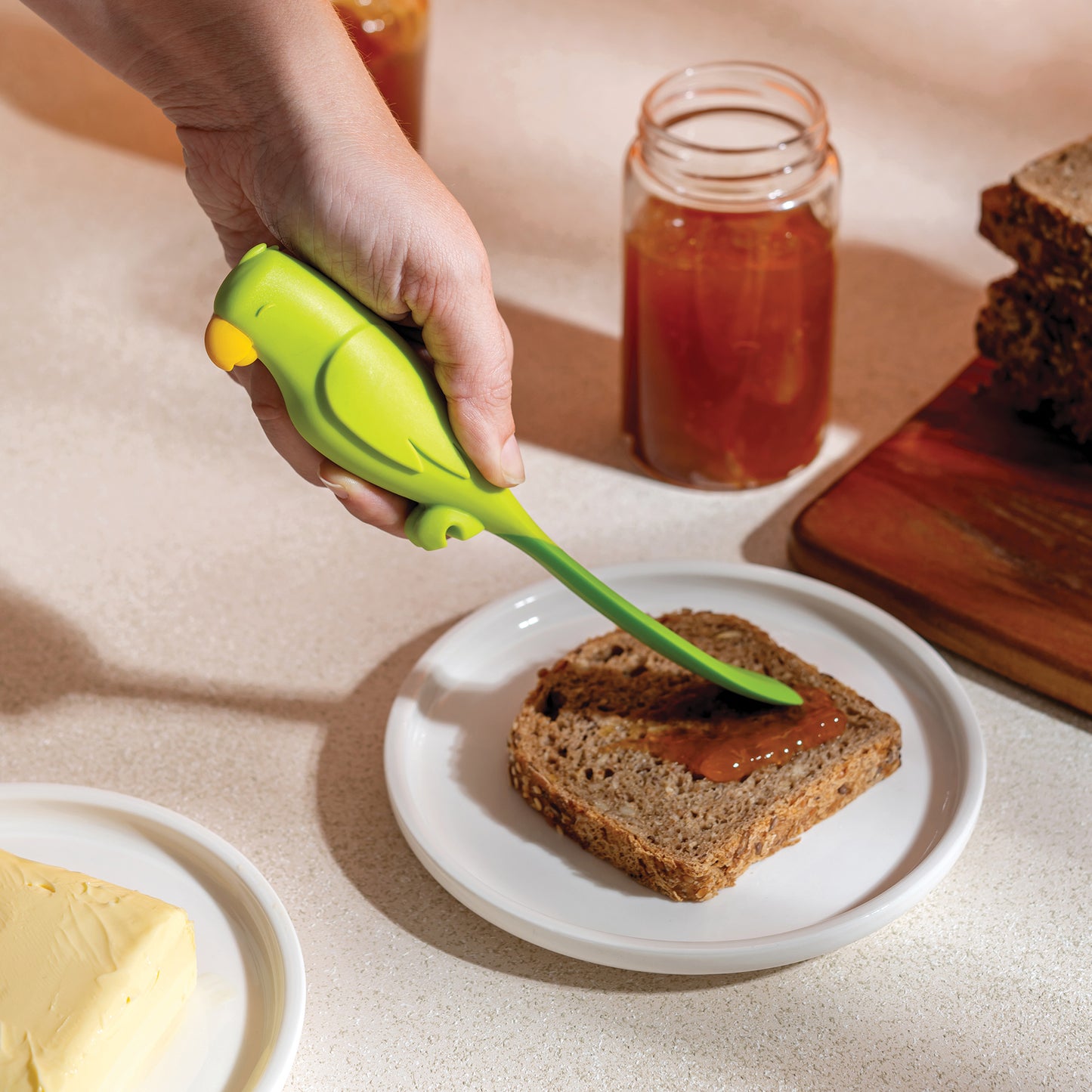 A hand uses the OTOTO Parry green bird-shaped spatula to spread jam on bread on a white plate.