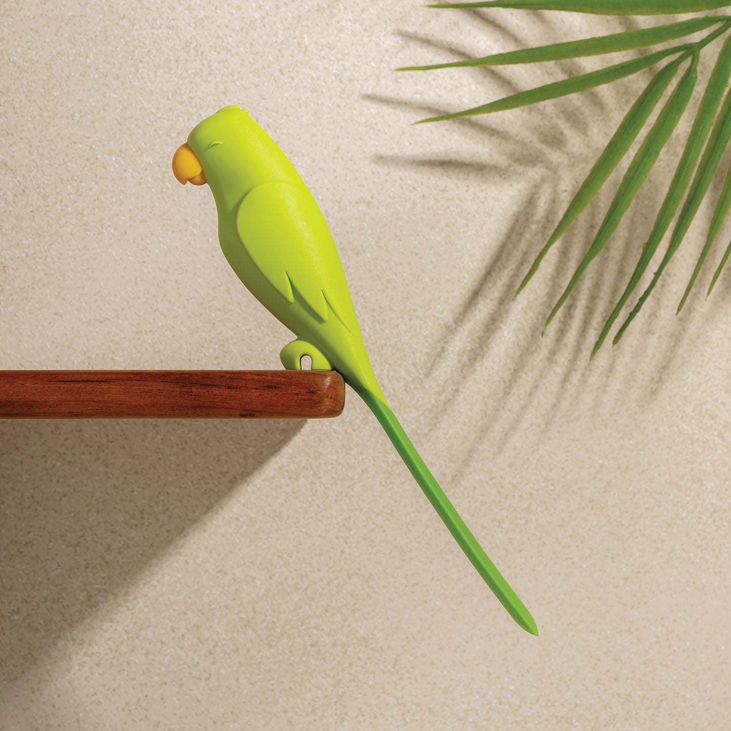 A green Parry bird figurine sits by an OTOTO silicone spatula on a shelf near a palm leaf shadow.