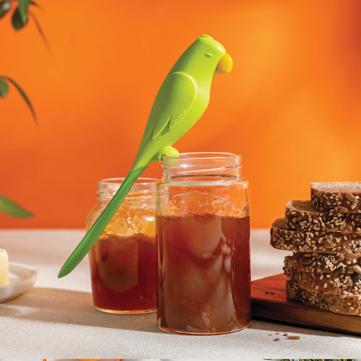 An OTOTO Parry green bird-shaped silicone spatula rests on a jam jar beside bread and another jar.