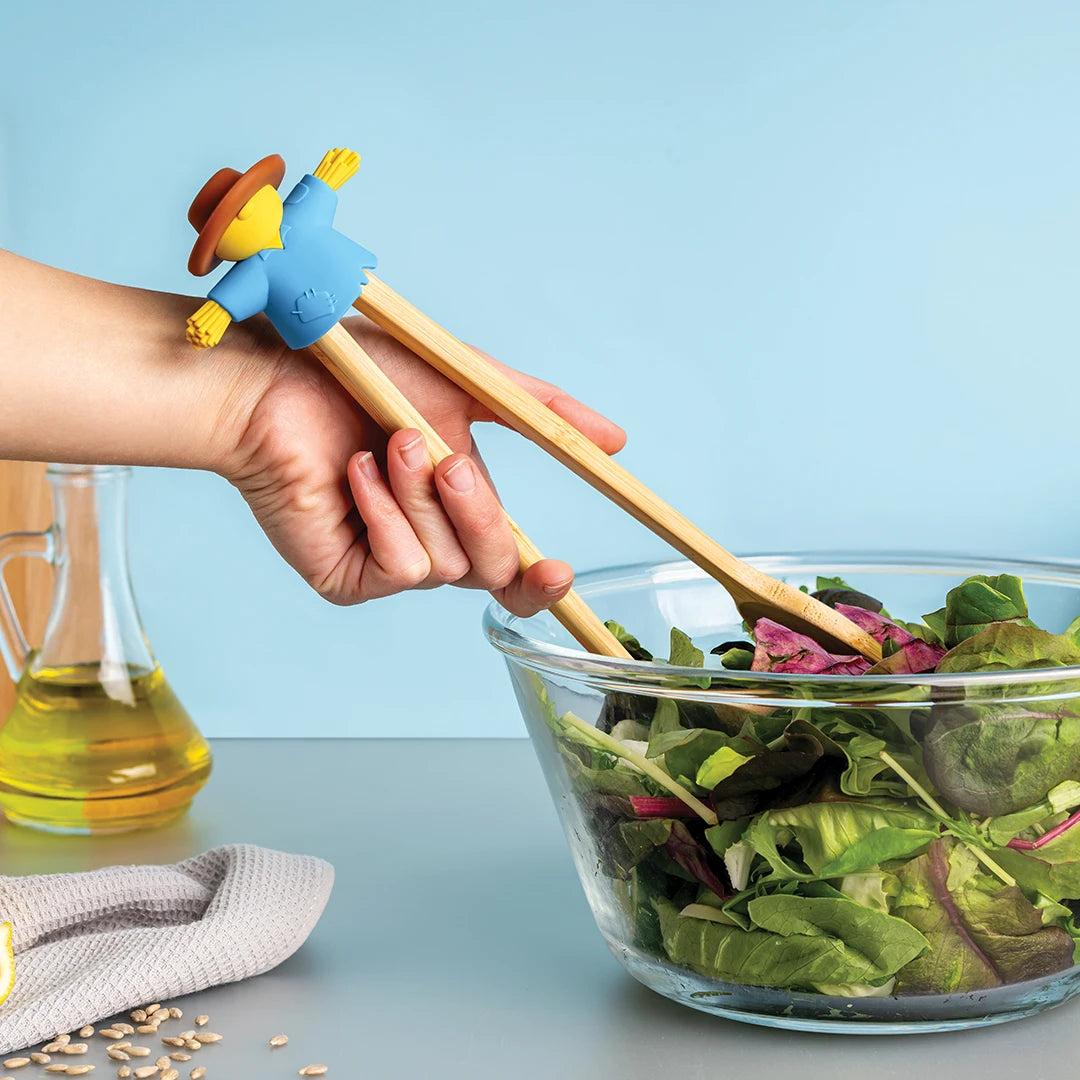 A hand uses OTOTOs Sal scarecrow tongs to toss a green salad in a glass bowl.